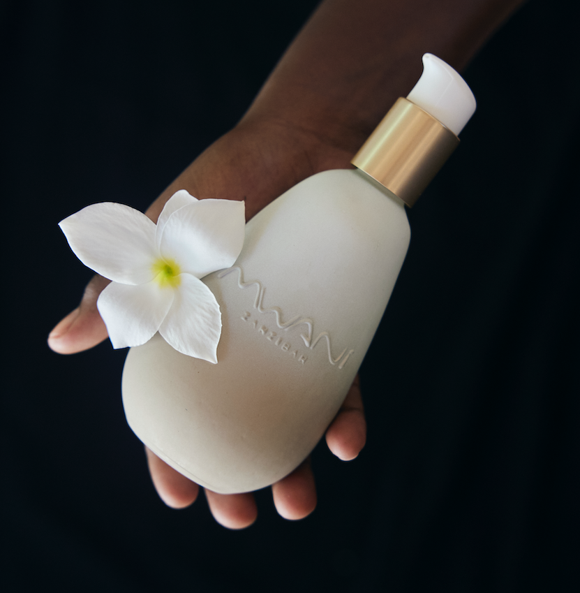 Hand holding a white and golden Mwani Zanzibar bottle and white flower with black background.
